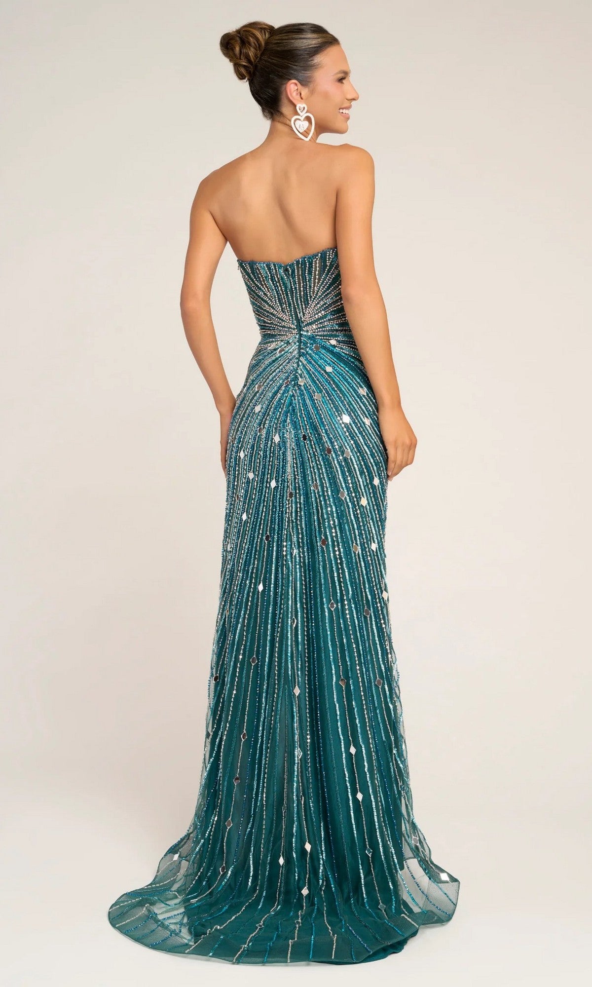 A woman stands with her back to the camera in a Portia and Scarlett PS26409C long formal dress—an elegant, strapless, floor-length teal gown adorned with sparkling beads and sequins. Her hair is in a bun, and she wears large heart-shaped earrings.